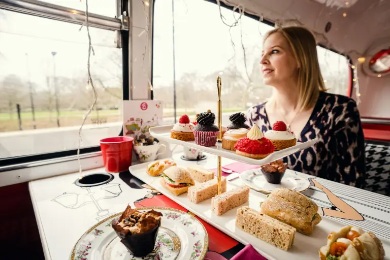 A woman enjoying a delightful afternoon tea on a scenic bus ride, celebrating Mother's Day with an elegant selection of pastries, sandwiches, and desserts. The cozy and charming setting highlights a unique and memorable way to honour mothers.