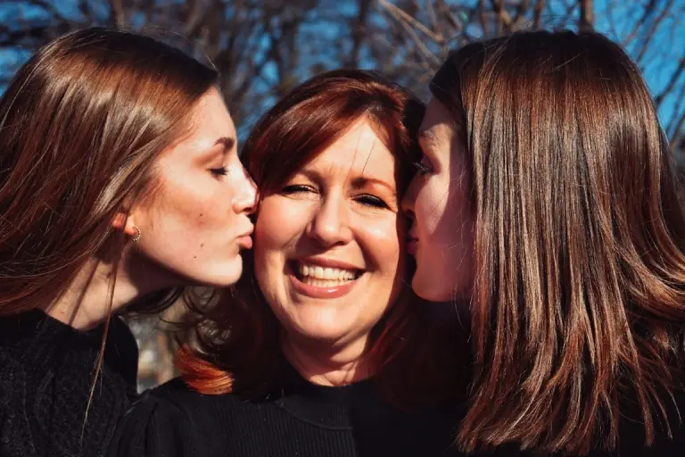 A mother joyfully smiling as her two daughters give her a loving kiss on the cheeks, capturing a heartfelt Mother's Day moment. This touching scene symbolizes appreciation, love, and the special bond between a mother and her children.